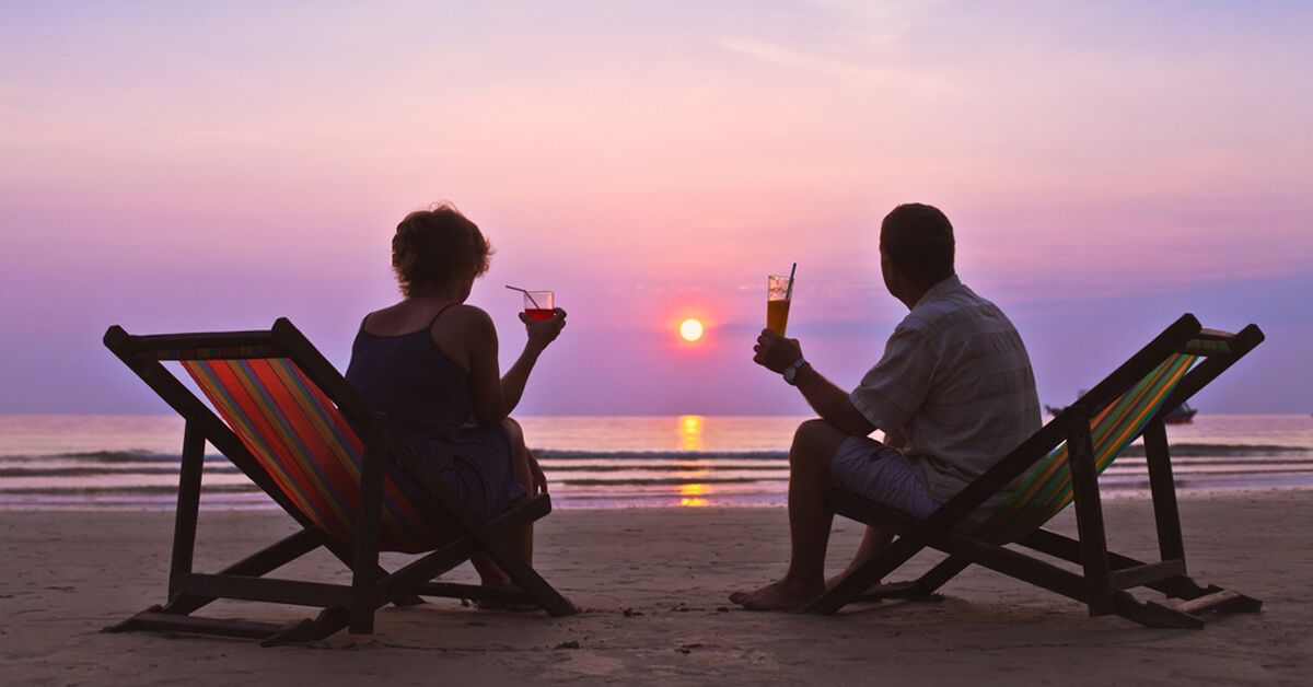 retired couple sits in chairs on the beach with drinks and watches sun set